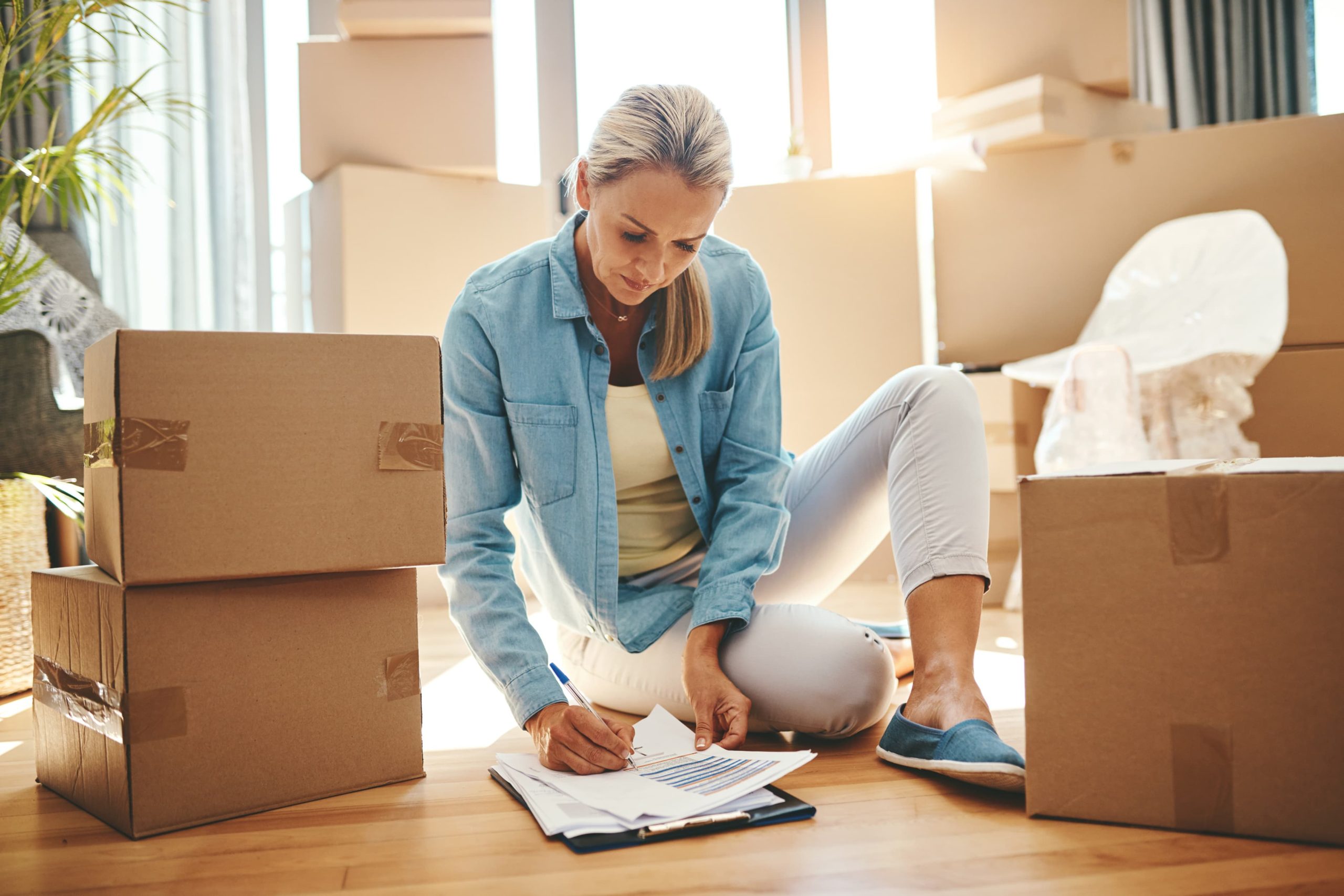 Mujer sentada en el suelo con la habitación llena de cajas y tomando notas.